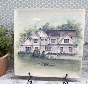 May include: Square ceramic plate featuring a hand-painted illustration of a house with a dark gray roof and white siding. The plate has a green and blue background with the text "The Bilsteins" in black.