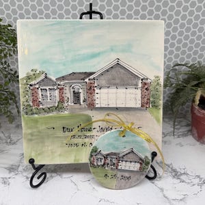 May include: A ceramic tile and ornament set featuring a watercolor painting of a house. The tile is square and mounted on a black stand. The ornament is round and has a gold ribbon. The house has a gray roof, white garage door, and brick accents.