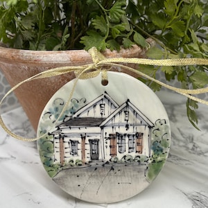May include: A round, white ceramic ornament featuring a hand-painted illustration of a house with a black roof, white siding, and brown shutters. The ornament is tied with a gold ribbon and has a hole at the top for hanging. The background includes greenery.