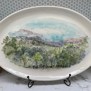 May include: White oval ceramic serving platter with a watercolor painting of a landscape scene. The painting depicts a green hillside with trees and a blue sky with white clouds. The text "The Highlands Holts" is written in black ink at the bottom of the platter.