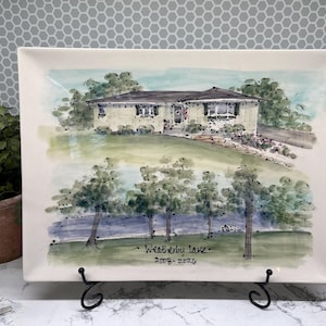 May include: A rectangular white ceramic platter featuring a watercolor-style illustration of a house with a lawn, trees, and a lake. The text "Weatherby Lake" and "2008-2024" is written in black ink at the bottom center of the painted scene.