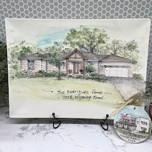 May include: Watercolor painting of a house with a garage, trees, and a driveway. The text "The Rodriguez Home - 7328 Wyoming Street" is written below the house. A round ornament with a house illustration and the text "The Jackmans married march 11, 2023" is also present.