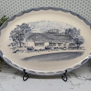 May include: An oval ceramic serving platter with a cream-colored base and a scalloped edge painted in blue. The center features a detailed blue ink-style illustration of a single-story house with trees, a front lawn, and a driveway.