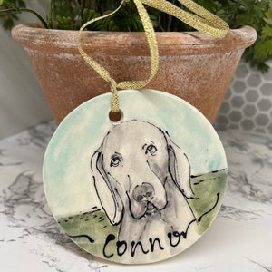 May include: A round ceramic ornament featuring a watercolor portrait of a dog with the name "Connor" in black script. The ornament has a light blue and green background and a gold ribbon for hanging.