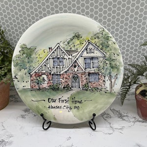May include: A decorative ceramic plate featuring a watercolor illustration of a house with a brick facade and a green lawn. The plate has the text "Our First Home - Kansas City, mo." displayed in a handwritten font.