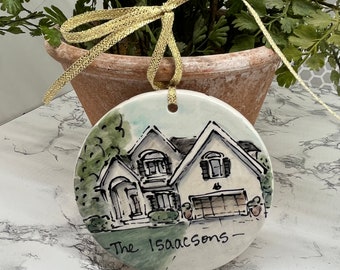 May include: A white ceramic ornament with a hand-painted illustration of a house and the text "The Isaacsons" on a green background. The ornament has a gold ribbon attached for hanging.
