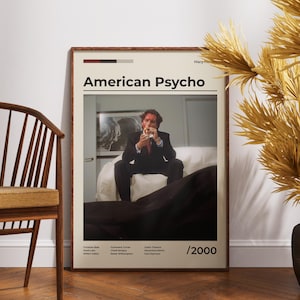 Könnte beinhalten: Ein gerahmtes Poster mit dem Titel "American Psycho" zeigt einen Mann im Anzug. Das Poster hat einen beigen Hintergrund mit schwarzem und rotem Text. Das Bild wird in einem Raum mit einem Holzstuhl und dekorativen Pflanzen angezeigt.