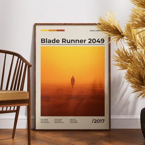Könnte beinhalten: Gerahmtes Poster für den Film "Blade Runner 2049" mit warmem, orange-gelbem Farbverlauf im Hintergrund. Das Poster zeigt eine einsame Figur, die auf den Betrachter zugeht. Das Poster befindet sich in einem Holzrahmen und wird neben einem Holzstuhl und dekorativen Trockenpflanzen ausgestellt.