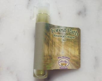 May include: A small glass bottle of Forest Path Botanical Cologne, labeled with a green and yellow image of a forest path. The label also includes the text "Forest Path Botanical Cologne 1ml Wise Mountain Botanicals".