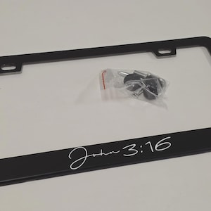 May include: Black license plate frame with the text "John 3:16" in white script. Includes mounting hardware in a small plastic bag. The frame is rectangular with rounded corners.