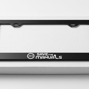 May include: A black rectangular license plate frame featuring a white graphic of a manual transmission gear shift pattern and the text SAVE THE MANUALS printed in white lettering along the bottom edge of the frame.