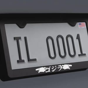May include: Black license plate frame with an Illinois license plate. The plate reads "IL 0001" in black lettering. A small American flag sticker is in the upper right corner. The bottom of the frame has white Japanese text and a Godzilla graphic.