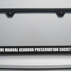 May include: Black license plate frame with the text "THE MANUAL GEARBOX PRESERVATION SOCIETY" in white. The frame has a rectangular shape with rounded corners and two small, round fasteners at the top.
