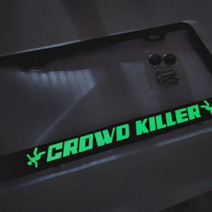 Puede incluir: Marco de matrícula negro con las palabras "CROWD KILLER" en verde brillante, junto con un gráfico verde de una persona. El marco incluye accesorios de montaje.