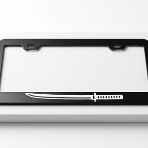 May include: A rectangular black license plate frame featuring a white graphic of a katana sword centered along the bottom edge. The frame has two mounting holes at the top and a smooth, matte finish, presenting a clean and simple design.