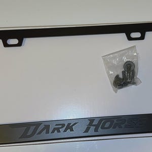 May include: Black license plate frame with the text "DARK HORSE" in a stylized font. The frame has a rectangular shape with rounded corners and mounting holes. A small bag of screws is included.