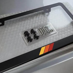 May include: Black license plate frame with a clear plastic cover. The frame has a color stripe of yellow, orange, and red. Includes mounting hardware.