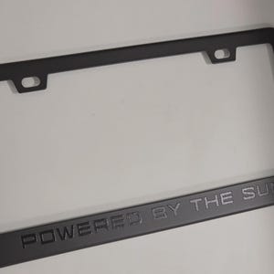 Puede incluir: Marco de matrícula negro con el texto en relieve "POWERED BY THE SUN". El marco tiene forma rectangular con esquinas redondeadas y orificios de montaje. El marco está hecho de un material negro mate.