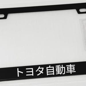 May include: Black license plate frame with the Japanese text "トヨタ自動車". Includes mounting hardware. The frame has a rectangular shape with rounded corners and a glossy finish. The frame is designed to fit a standard U.S. license plate.