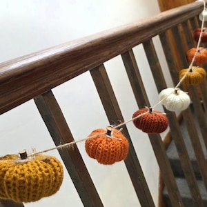 May include: A garland featuring multiple small, knitted pumpkins in shades of mustard yellow, burnt orange, and cream. Each pumpkin is attached to a thin, natural-coloured twine string and topped with a small, dark brown wooden stem.