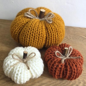 May include: Three decorative pumpkins crafted from knit yarn in mustard yellow, cream white, and burnt orange. Each pumpkin features a small wooden stick as a stem, accented with a tied piece of natural jute twine wrapped around the base of the stem.