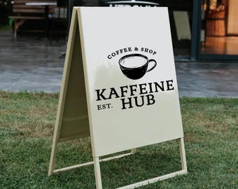 May include: A-frame sign with the text "Coffee & Shop" and "Kaffeine Hub Est." in black. The sign features a black coffee cup graphic and is set on a grassy area.