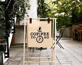 Puede incluir: Un letrero en forma de caballete con las palabras "Coffee To Go" en letras negras. El letrero presenta un sombrero de copa y un gráfico de taza de café encima de una rueda de monociclo. El letrero es de color crema y cuelga de un marco de metal claro con correas de cuero marrón.