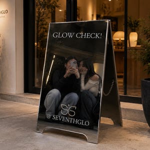 Puede incluir: Un letrero de espejo en forma de A plateado con una superficie reflectante que presenta el texto blanco GLOW CHECK! en la parte superior y el logotipo con el texto @SEVENTHGLO en la parte inferior. El espejo refleja a dos personas.