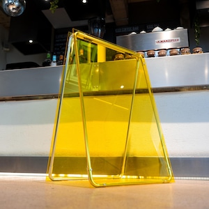 May include: A-frame sign with a transparent yellow panel and a gold-coloured metal frame. The sign is standing on a light-coloured surface, likely indoors. The sign is empty, ready for custom messaging.