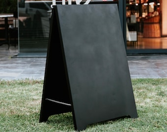 May include: A-frame chalkboard sign, painted black, standing on green grass. The sign is blank, ready for custom messages. The sign is positioned outdoors, likely for a business to display menus or promotions.