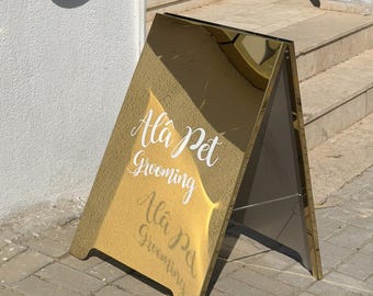 May include: A gold-colored A-frame sign with a reflective, mirror-like surface. The text "Alâ Pet Grooming" is printed in white script lettering on the front panel. The sign features a sturdy metal frame and a thin chain connecting the two sides.