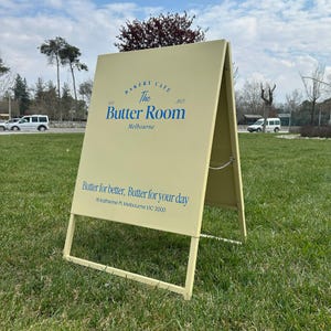 Op de afbeelding: Een lichtgeel A-framebord voor bakkerij en café "The Butter Room". Het bord heeft blauwe tekst met de naam, locatie en slogan "Butter for better, Butter for your day".