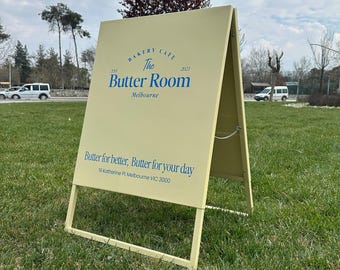 May include: A-frame sign for "The Butter Room" bakery and cafe. The sign is light yellow with blue text, including the bakery's name, location, and slogan "Butter for better, Butter for your day."