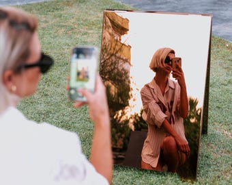 May include: A person taking a photo of a mirror with a phone. The mirror reflects a person in a striped shirt and sunglasses. The scene is set outdoors on a grassy area, with the sky visible in the background.