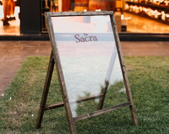 May include: A-frame sign with a clear glass panel and a dark wooden frame. The sign displays the word "Sacra" in a burgundy font. The sign is positioned on a grassy area, with a blurred background.