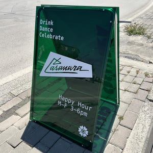 Könnte beinhalten: Ein grünes, doppelseitiges Werbeschild mit den Worten "Drink Dance Celebrate" oben. Das Schild zeigt das Logo "Casanara" und den Text "Happy Hour M-F 3-6pm". Eine weiße Blumengrafik befindet sich unten.