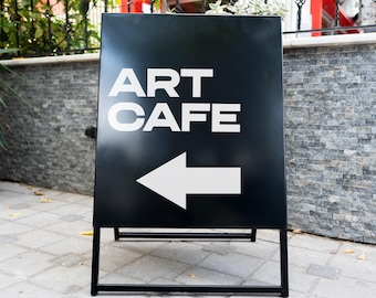 May include: A black metal sandwich board sign with the words "ART CAFE" in white, and a white arrow pointing left. The sign is on a black metal frame, set on a brick and stone paved surface.
