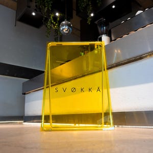 May include: A bright yellow, transparent sign with the word "SVØKKÄ" in black lettering. The sign is in a frame and stands on a light-colored surface. The background includes a counter, plants, and a disco ball.