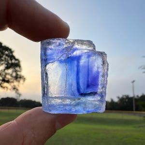 Könnte beinhalten: Ein rechteckiger, durchscheinender Kristall mit Blau- und Lilatönen. Der Kristall wird vor dem Hintergrund einer grünen Wiese und eines sanften, sonnenuntergangsfarbenen Himmels gehalten. Die Kanten des Kristalls sind rau und uneben.