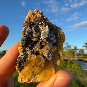 Pode incluir: Um espécime mineral natural bruto com uma mistura de cores amarelo, castanho e cinza escuro. A rocha tem uma superfície texturizada e parece estar sendo segurada em uma mão, com um céu azul e grama verde ao fundo.