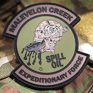 May include: A circular morale patch with a sage green background and black border. It features a pink skull biting a black knife dripping oil. The text reads MALEVELON CREEK at the top and EXPEDITIONARY FORCE at the bottom, with SPILL OIL in the center.