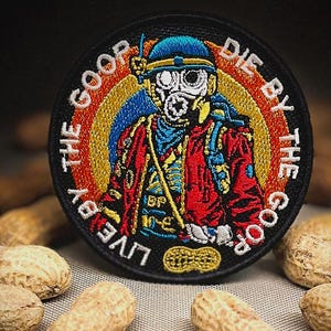May include: Embroidered patch with a circular design featuring a figure in a gas mask and military gear. The patch has the text "LIVE BY THE GOOP DIE BY THE GOOP" in a circular pattern. The patch is surrounded by peanuts.