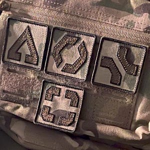 May include: A camouflage tactical pouch with four embroidered patches. The patches feature geometric designs in shades of brown and tan. The pouch is made of a durable, textured fabric and has a zipper closure.