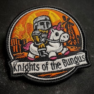 May include: Embroidered patch featuring a knight riding a unicorn with a pink mane and hooves. The knight is wearing armor and holding a weapon. The patch has the text "Knights of the Bungus" on a banner.