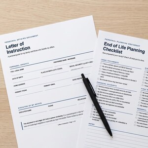 May include: Two white printed documents titled Letter of Instruction and End of Life Planning Checklist with black text and blue lines, accompanied by a black ballpoint pen resting diagonally across the pages on a light wood surface.