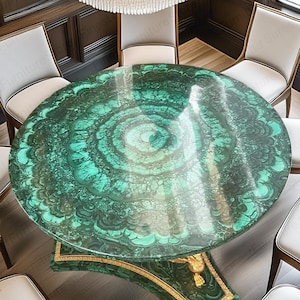 Può includere: Un tavolo da pranzo rotondo con piano verde malachite e base con finiture dorate. Diverse sedie con cuscini bianchi e struttura in legno circondano il tavolo. Un grande lampadario è appeso sopra il tavolo, illuminando la stanza.
