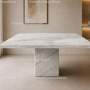 Puede incluir: Una gran mesa de comedor cuadrada de mármol con una sola base rectangular. La encimera presenta un patrón veteado gris y blanco. La mesa está en una habitación de tonos neutros con una ventana y obras de arte.