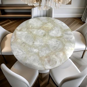 May include: A round dining table with a light-colored, patterned surface, surrounded by four white chairs. The table's surface has a unique, natural stone appearance. The text "FinestFurnitureStore" is visible on the table. The chairs have a modern design.