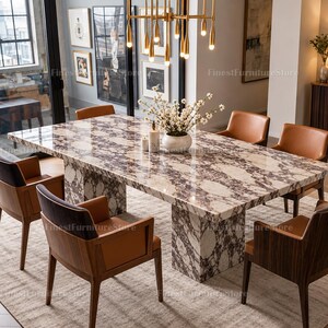 May include: A rectangular dining table with a white and purple marble top and base, surrounded by six wooden armchairs with brown leather upholstery. A gold multi-light pendant chandelier hangs above, and a white vase with flowers sits on the table.