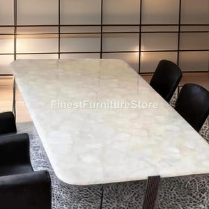 May include: A rectangular marble-topped dining table with rounded corners. The table has dark brown legs and is surrounded by black chairs. The text "FinestFurnitureStore" is visible on the table's surface. The table is set on a patterned rug.